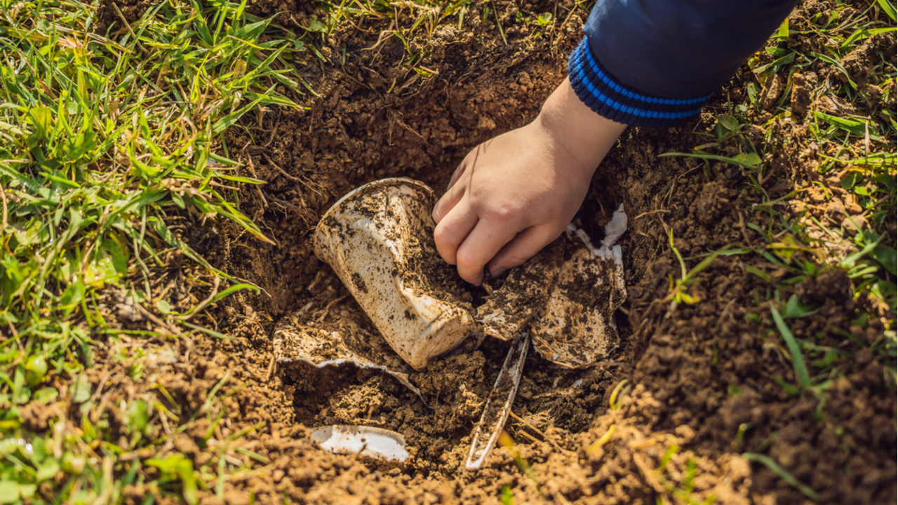 En esta imagen vemos una mano enterrando plásticos biodegradables de un solo. 