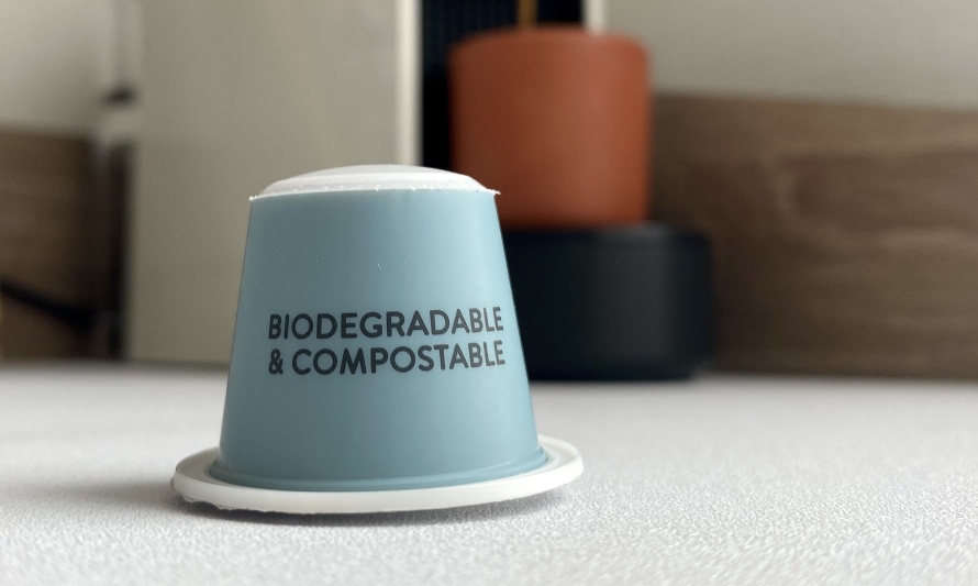 En esta imagen vemos una cápsula de café que tiene impresas las palabras biodegradables y compostable. Esta cápsula es de color azul pálido y está sobre una mesa.